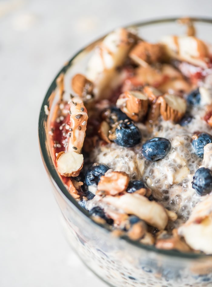 Close up of a bowl of overnight chia seed pudding with oats topped with almonds, blueberries, jam and sliced banana. 