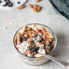 A bowl of blueberry and banana chia seed oatmeal topped with nuts and sliced banana. There is a spoon in the bowl.