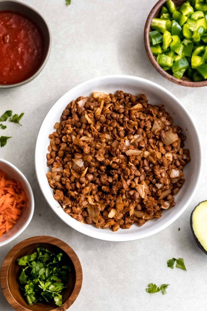 Vegan lentil taco filling in a white bowl plus bowls with carrot, avocado, salsa and cilantro.