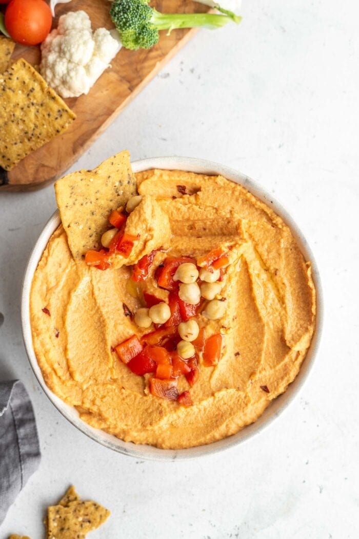 Bowl of vegan roasted red pepper hummus with a tortilla chip being dipped in it.