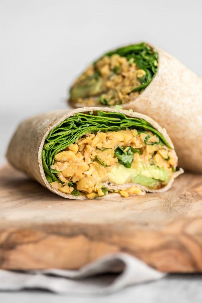 Close up of a spicy chickpea salad wrap with spinach and avocado cut in half on a cutting board.