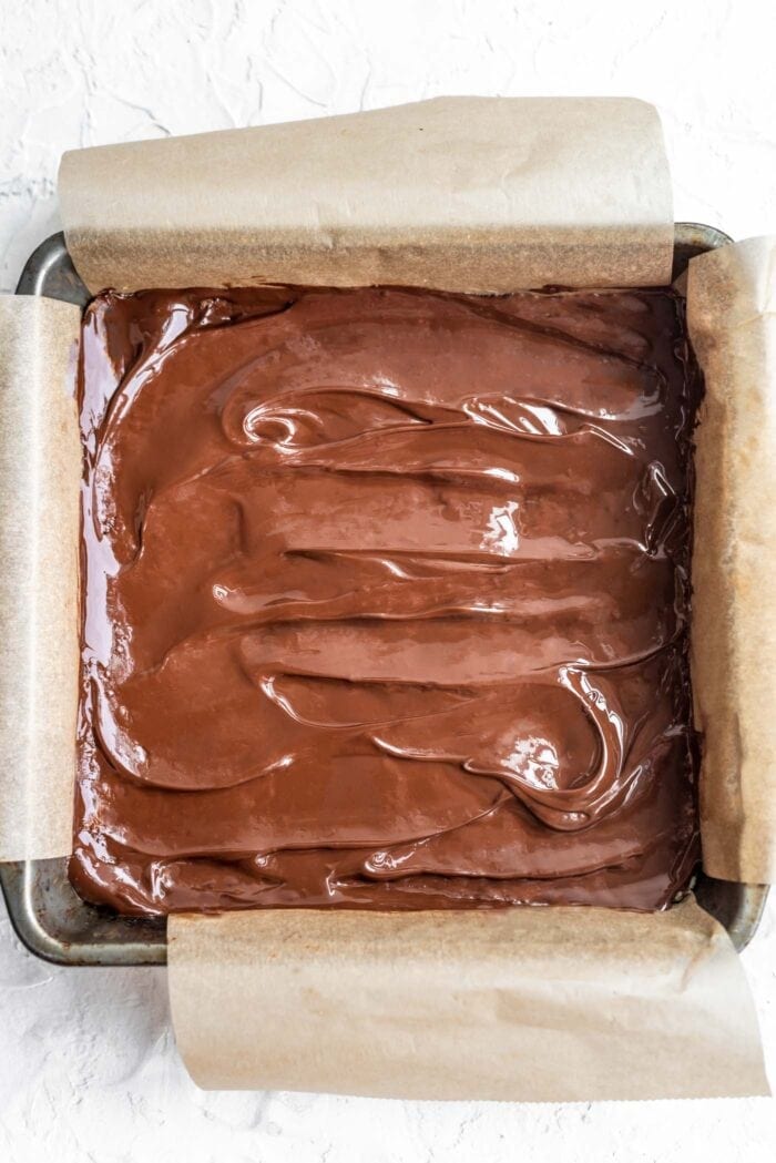 Melted dairy-free chocolate spread over coconut bars in a baking pan lined with parchment paper.