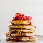 A stack of berry banana pancakes topped with chopped strawberries being drizzled with maple syrup.
