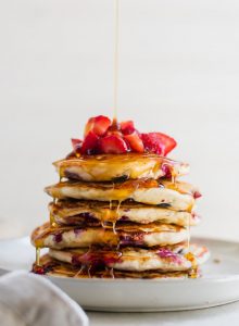 A stack of berry banana pancakes topped with chopped strawberries being drizzled with maple syrup.