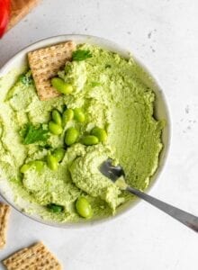 A spoon and cracker scooping edamame hummus out of a small bowl sitting on a white surface.