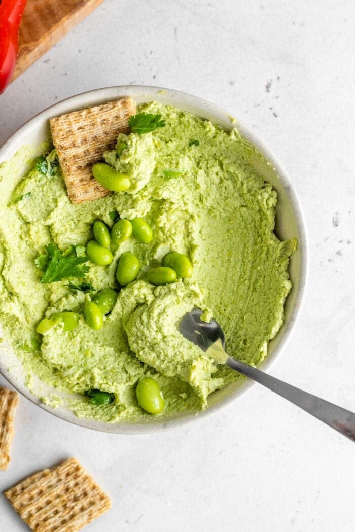 A bowl of edamame hummus topped with a few pieces of cilantro and edamame beans with a cracker and spoon in the bowl.