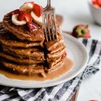 Fork sticking into a stack of vegan peanut butter banana pancakes with strawberries and peanut butter sauce.