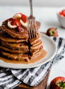 Fork sticking into a stack of vegan peanut butter banana pancakes with strawberries and peanut butter sauce.