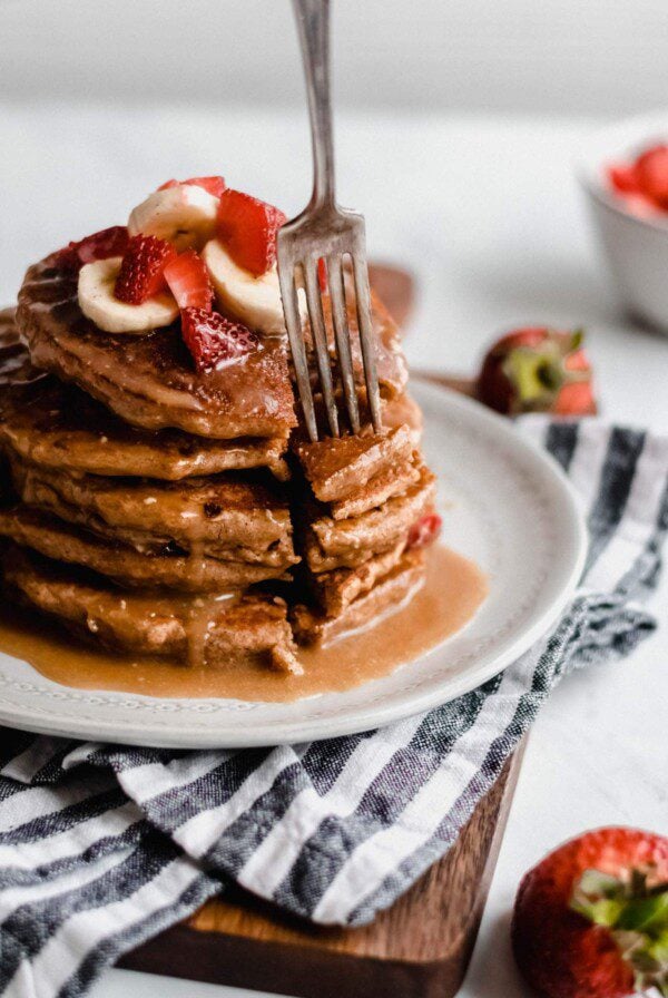 Fork sticking into a stack of vegan peanut butter banana pancakes with strawberries and peanut butter sauce.