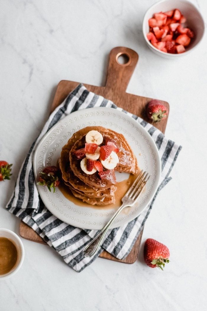 Overhead view of a stack of strawberry peanut butter banana pancakes on a plate on a cutting board topped with fresh berries, banana and syrup.