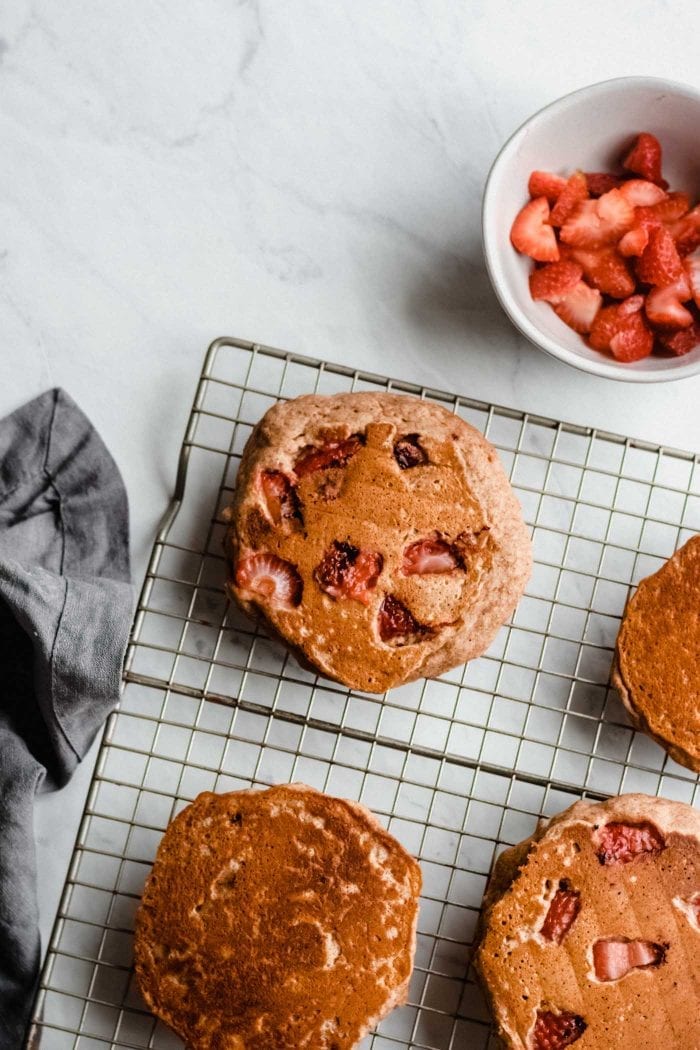 Peanut butter banana pancakes with strawberries sitting on a baking cooling rack. A small bowl of chopped strawberries is beside the rack.