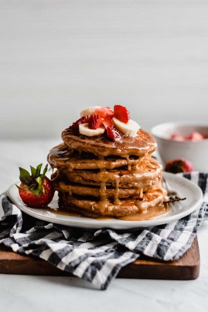 Stack of peanut butter banana pancakes topped with sliced banana, strawberries and peanut butter sauce on a small plate with a fresh strawberries resting on the plate..