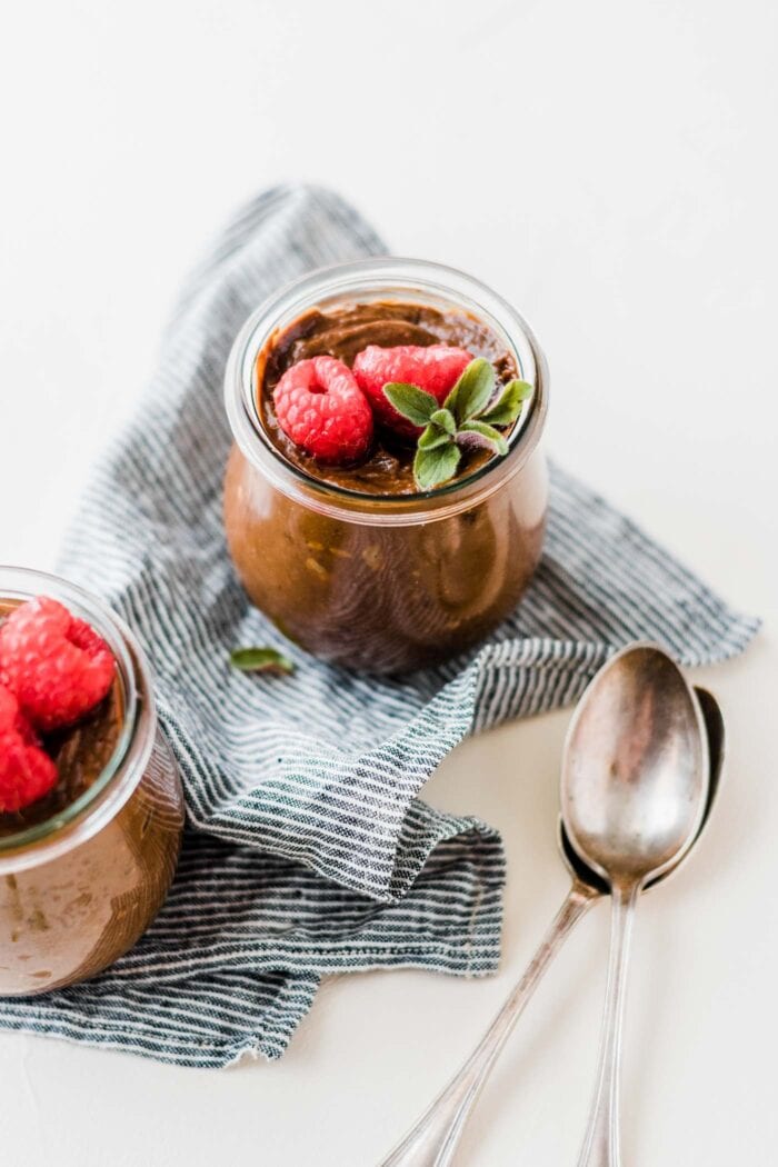 Two small glass jars of chocolate avocado pudding sitting on a striped napkin on a white surface.