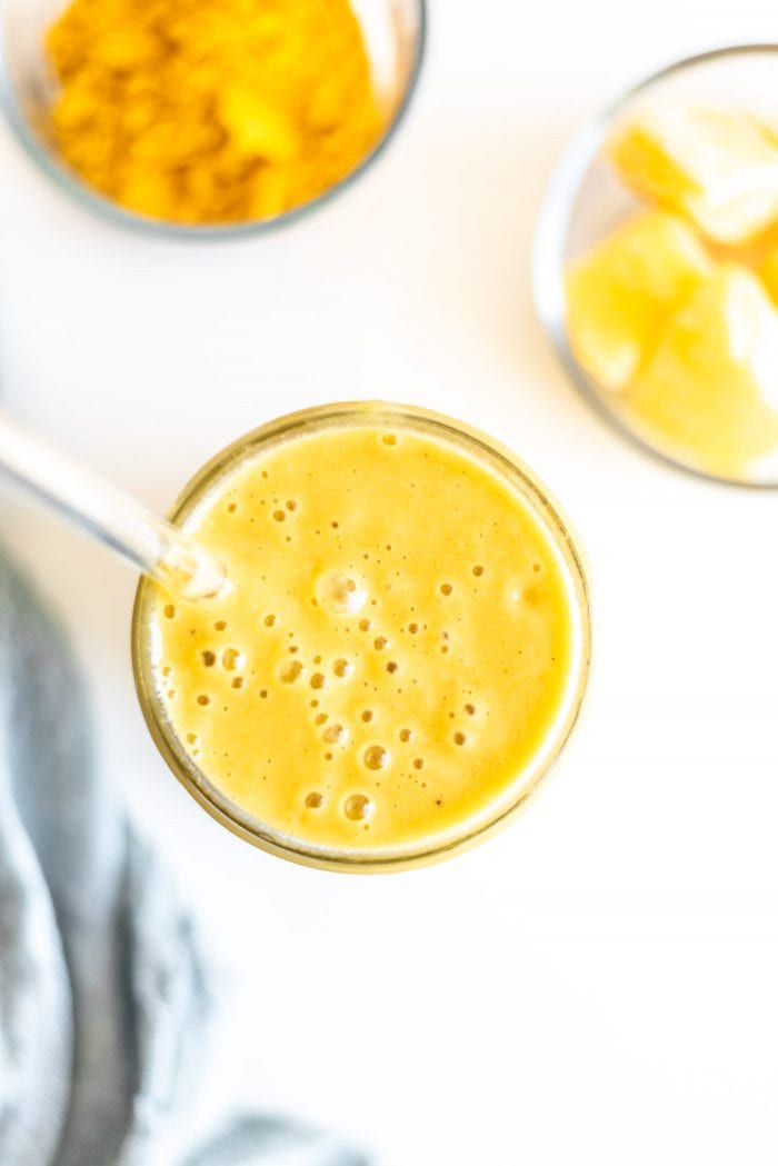An orange mango smoothie in a glass jar with a dish of turmeric and frozen mango.
