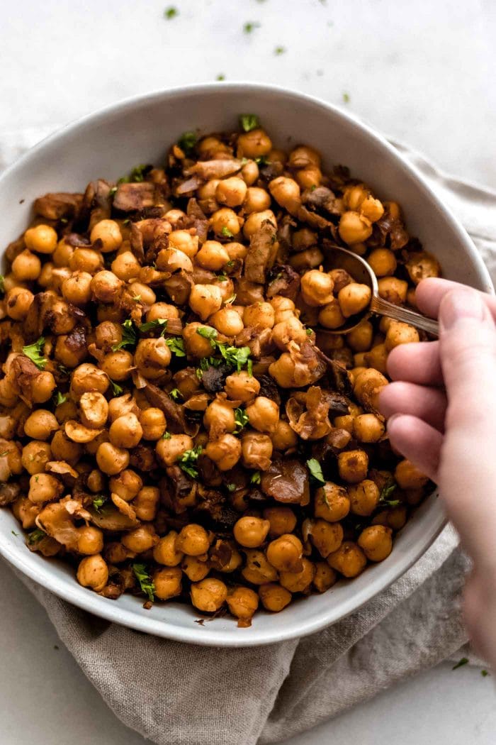 Hand using a spoon to serve some vegan spiced chickpeas with mushrooms and garlic.