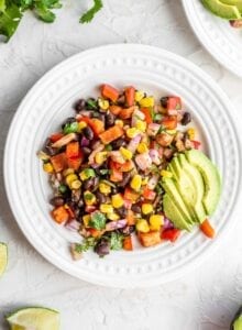 Black bean corn salad with avocado on a small white plate with cilantro and lime on the side.