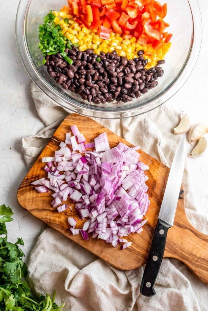 Chopped red onion on a cutting board beside a bowl of black beans, corn and bell pepper.