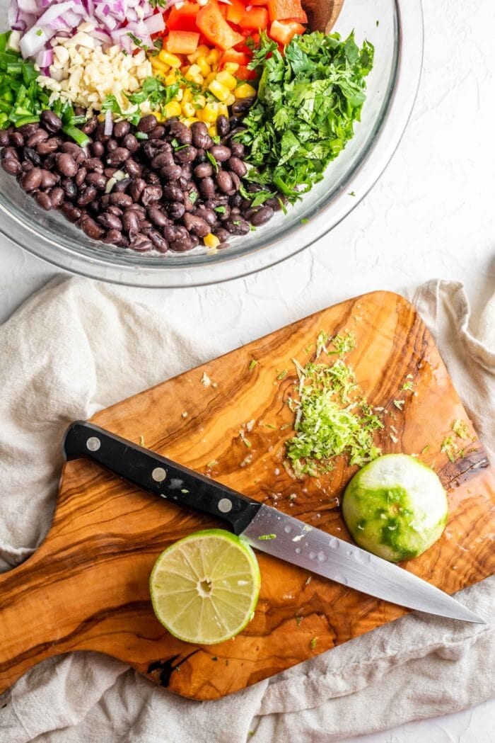 A knife chopping lime zest on a cutting board beside a bowl of black bean corn salad.