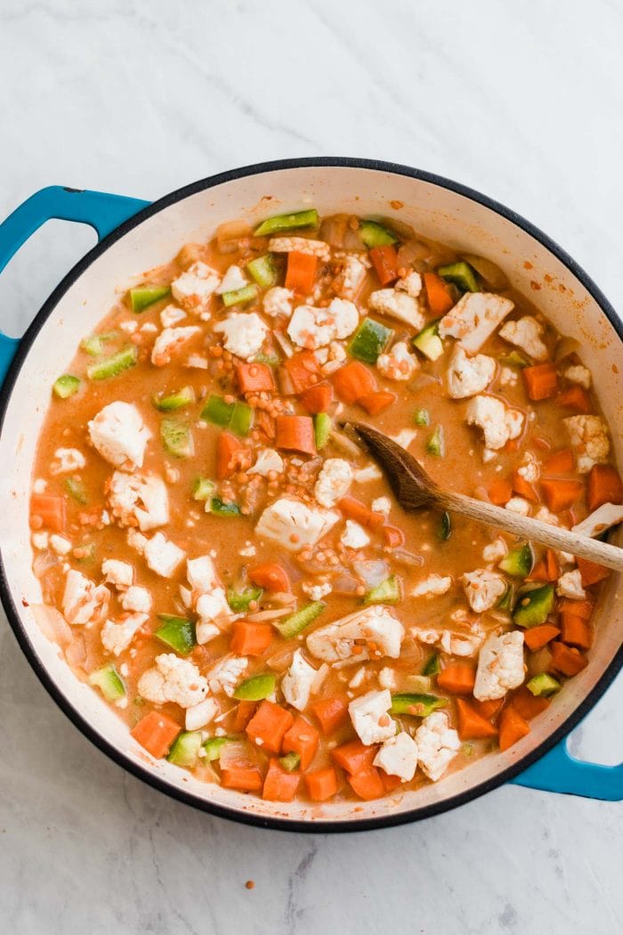 Carrot, cauliflower, celery and onion cooking in a red curry broth in a large pot. A wooden spoon rests in the pot.