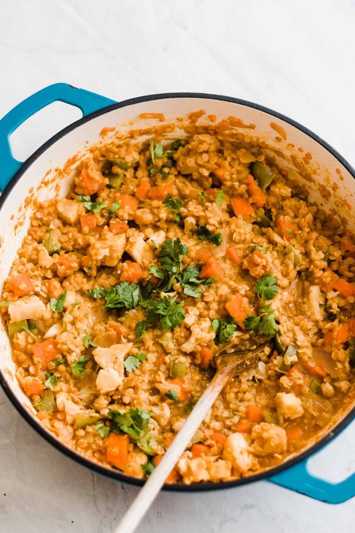 A red lentil curry dish with various vegetables cooking in a large pot with a wooden spoon.