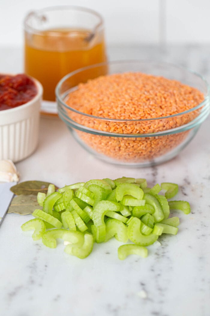 Chopped celery on a marble counter top. There are bowls of red lentils, vegetable broth and diced tomatoes in the background.