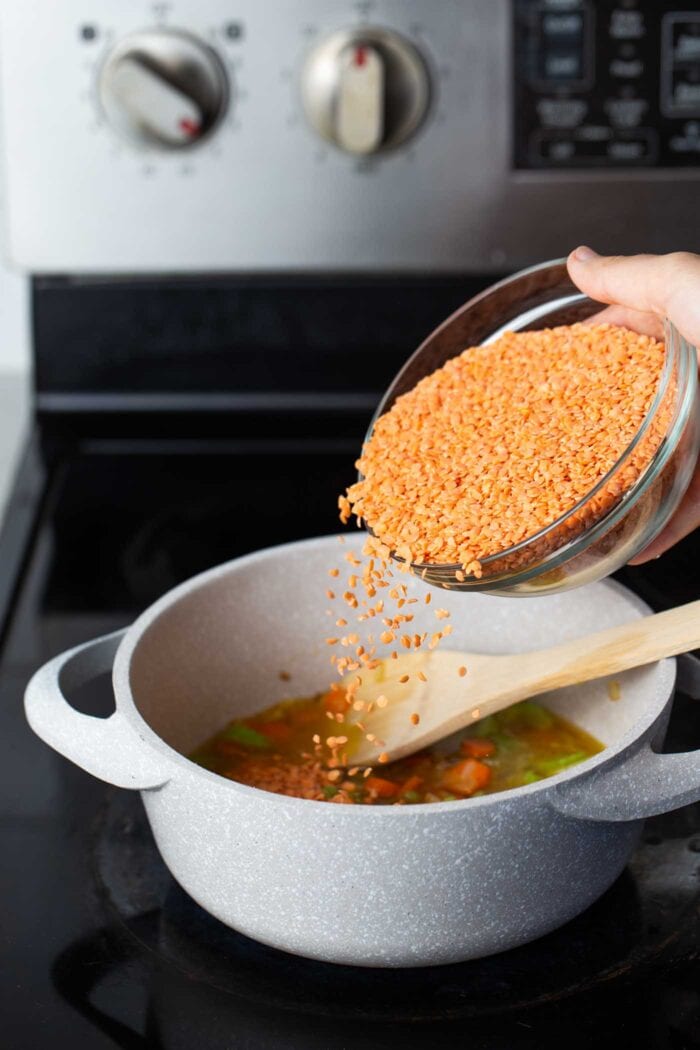 A hand pouring a bowl of red lentils into a soup pot on the stovetop.