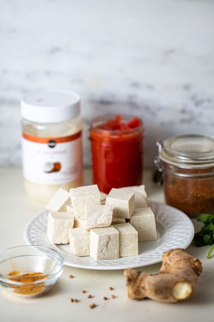 Cubed tofu on a small white plate with a piece of raw ginger and small bowl of spices beside it.