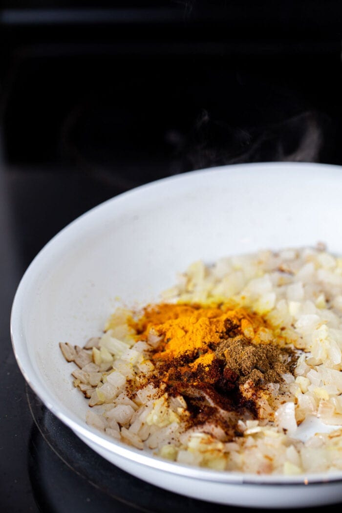 Indian spices like turmeric, cumin and chili powder being added to sauteed onions, garlic and ginger in a skillet.