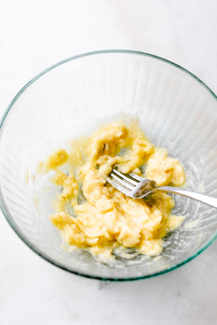 Mashed banana with a fork in a glass bowl.