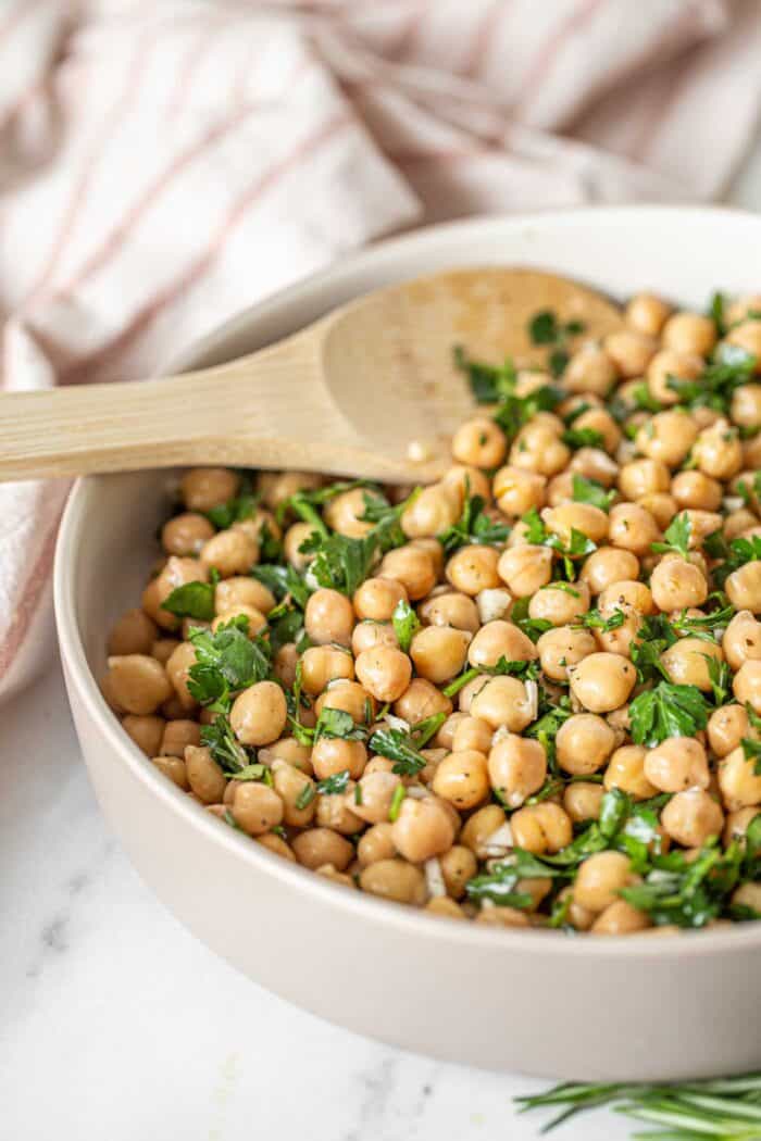 A serving spoon in a bowl of chickpeas with chopped herbs and garlic.