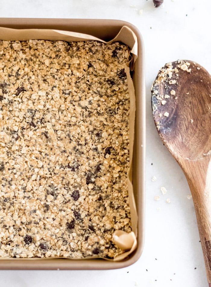 Chocolate chip oatmeal dough firmly pressed into a lined square baking pan. Wooden spoon rests beside.