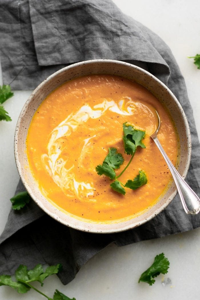 Overhead view of a bowl of creamy carrot ginger soup topped with a drizzle of coconut and a few leaves of cilantro. A spoon rests in the bowl.