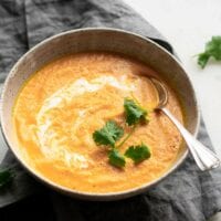 An angled view of a bowl of creamy carrot ginger soup sitting on a folded dish cloth and topped with a drizzle of coconut and a few leaves of cilantro. A spoon rests in the bowl.