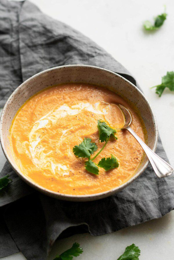 An angled view of a bowl of creamy carrot ginger soup sitting on a folded dish cloth and topped with a drizzle of coconut and a few leaves of cilantro. A spoon rests in the bowl.