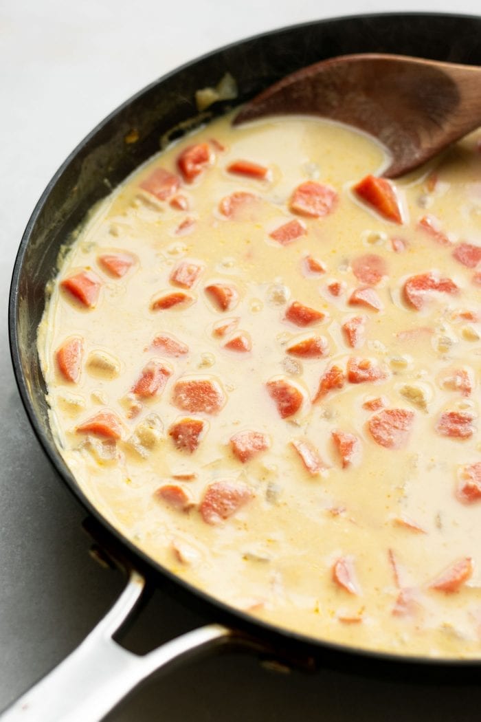 A creamy soup with carrot chunks cooking in a skillet with a wooden spoon in it.