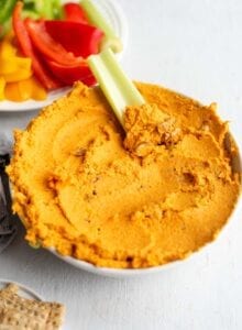 A bowl of roasted carrot hummus with a piece of celery dipped in it.