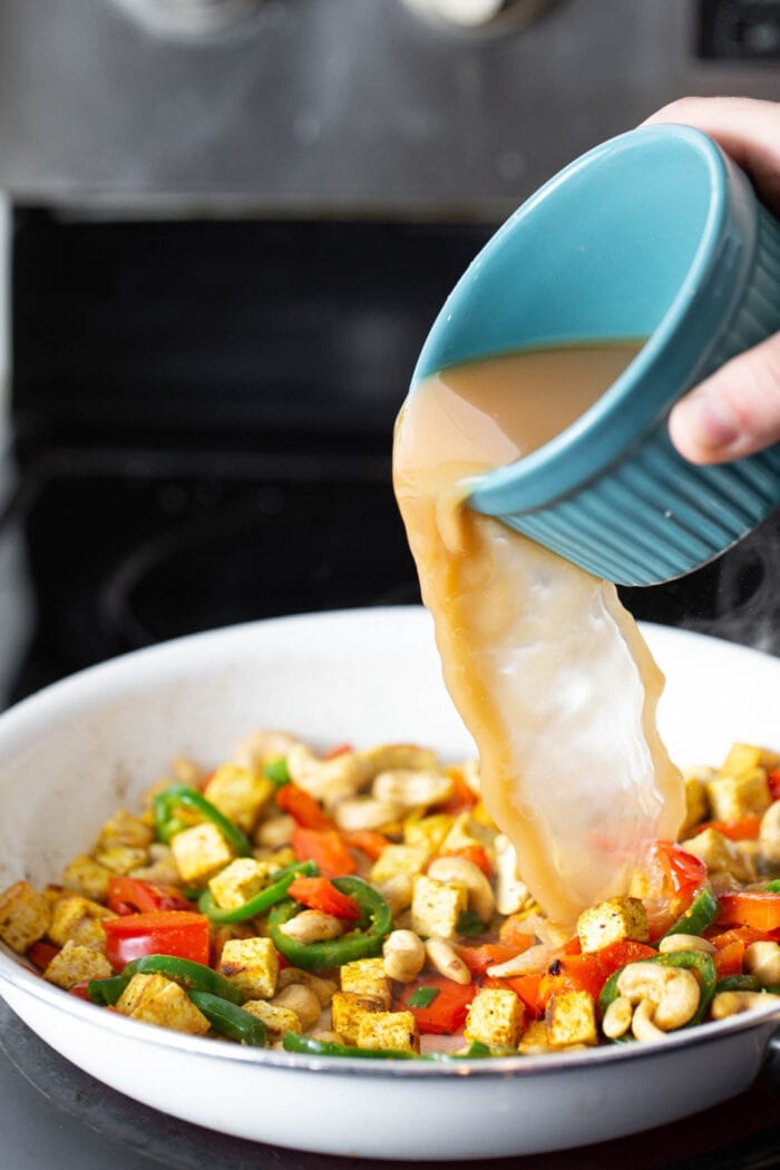 Vegetable broth being poured into a skillet of tofu, cashews and veggies on the stovetop.
