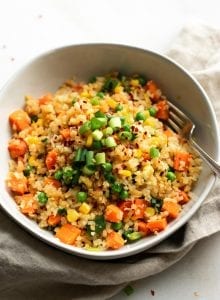 A bowl of fried cauliflower rice with carrots, peas and corn, topped with green onion with a fork in the bowl.