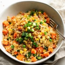 A bowl of fried cauliflower rice with carrots, peas and corn, topped with green onion with a fork in the bowl.