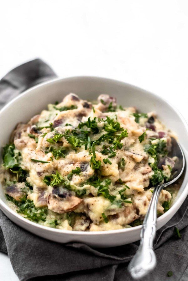 Close up of a bowl of mashed cauliflower with kale and mushrooms sitting on a grey napkin on a white surface.