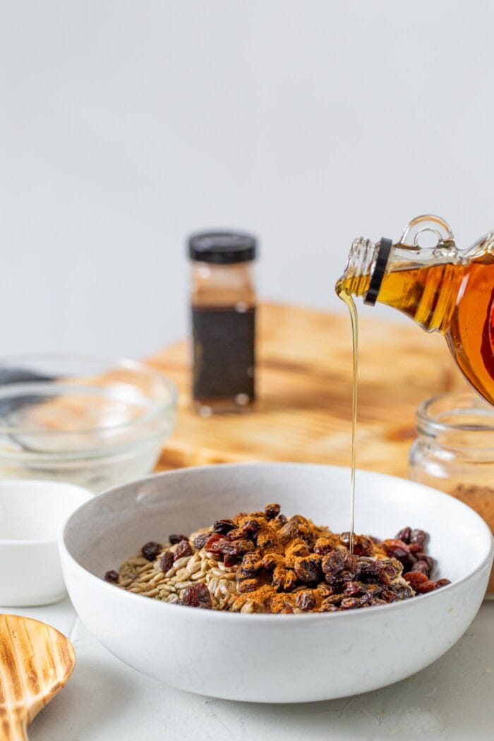 Maple syrup being poured into a mixing bowl of oats, cinnamon and raisin.