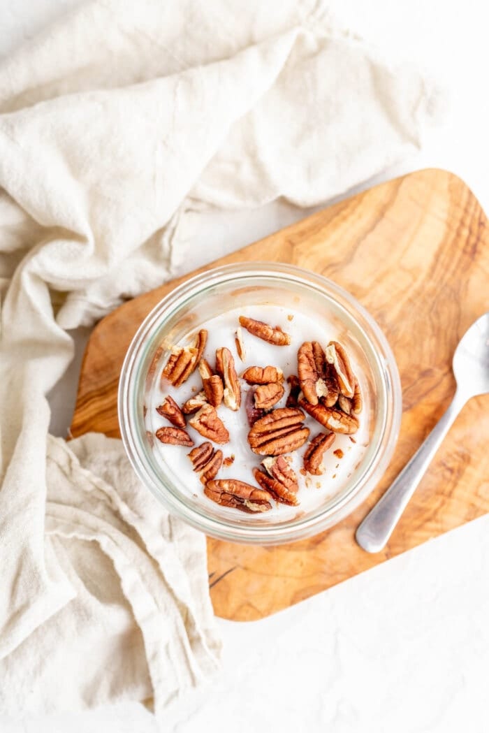 Overhead image of a jar of coconut yogurt and pecans sitting on a cutting board with a spoon beside it.