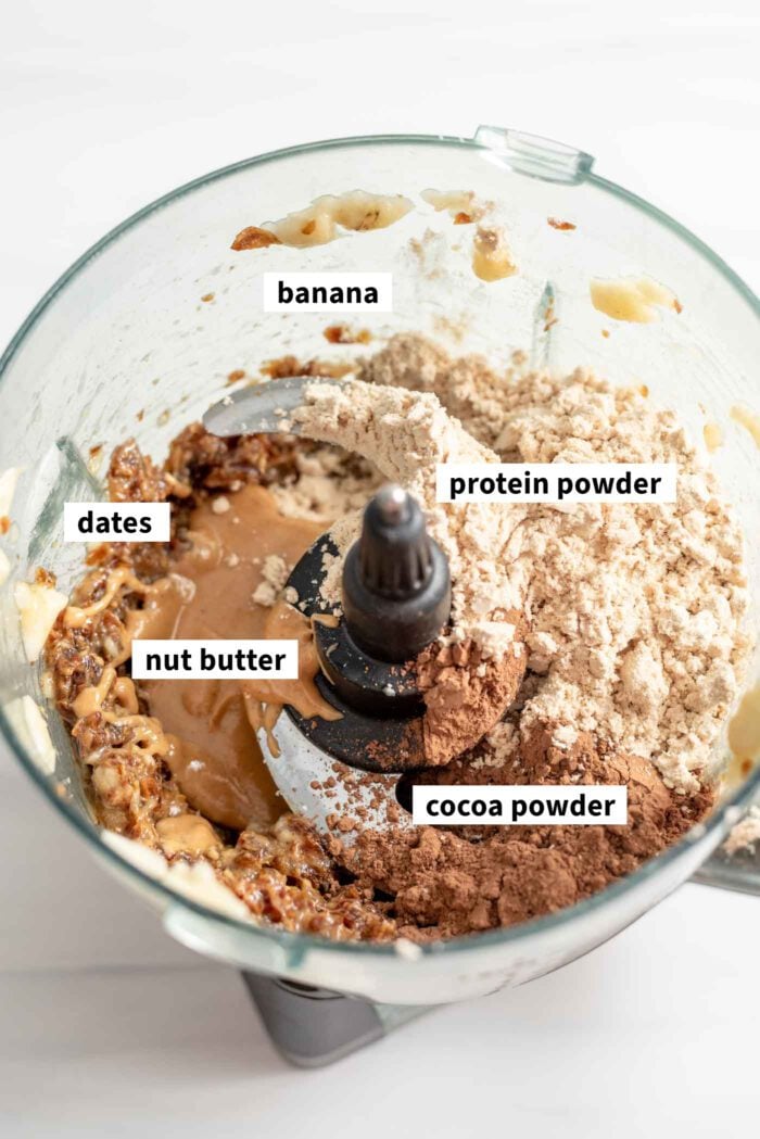 Protein powder, cocoa powder, dates, banana and peanut butter in a food processor. Each ingredient is labelled.