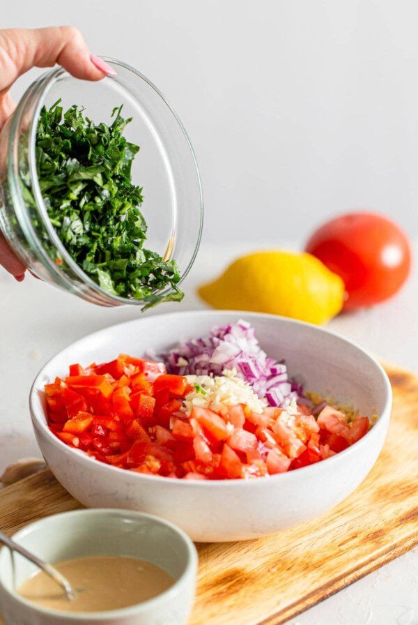 A bowl of chopped parsley being added to a bowl of fresh chopped veggies.