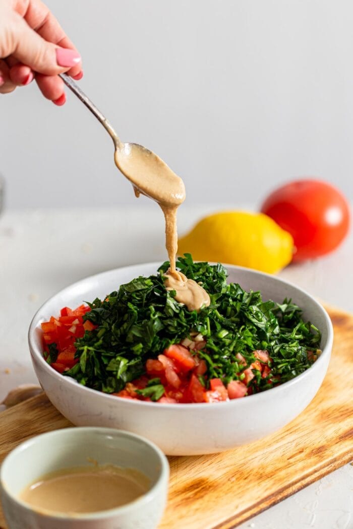 A spoonful of tahini being added to a bowl of fresh chopped parsley and bell pepper.