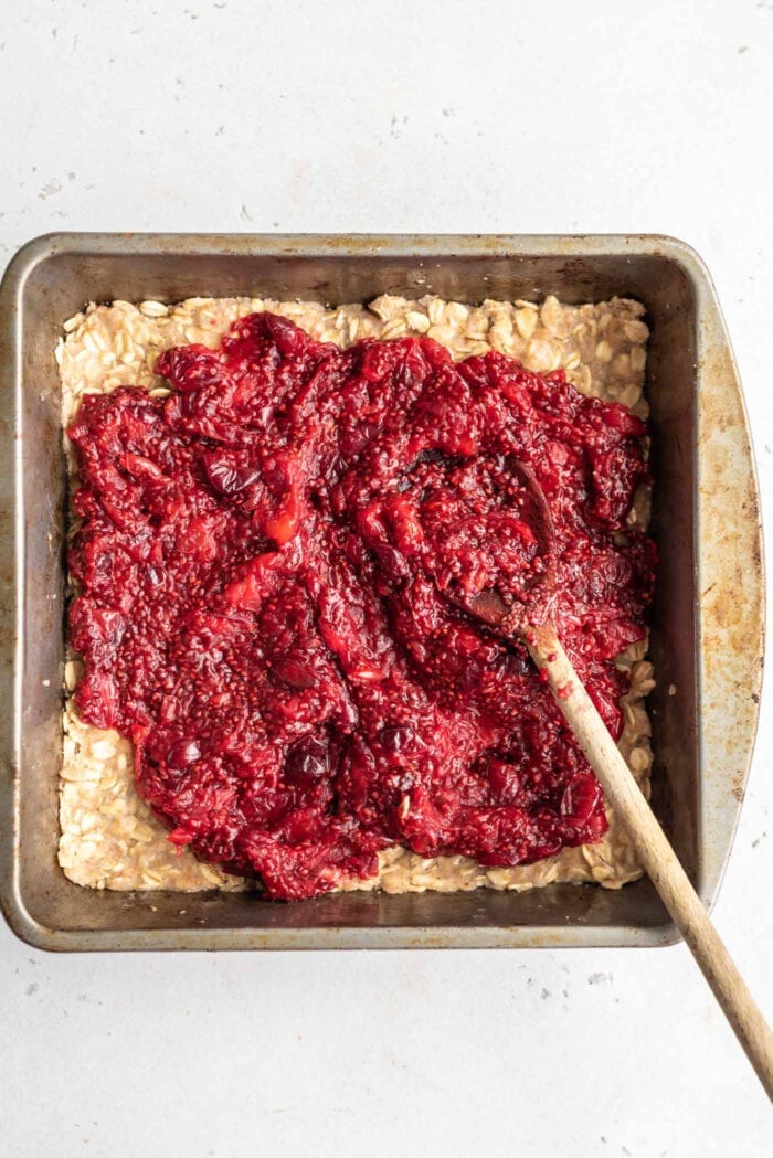 Cranberry jam being spread on a layer of oat crumble dough in a baking pan.