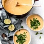 Two bowls and a large stock pot of cauliflower sweet potato soup with green peas. The bowls of soup are topped with fresh cilantro and there are some wedges of lime and two spoons beside them.