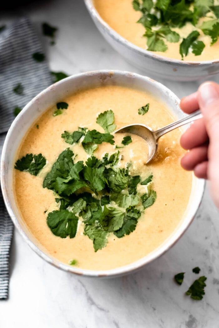 A hand stirring a bowl of creamy cauliflower sweet potato soup topped with fresh cilantro with a spoon.