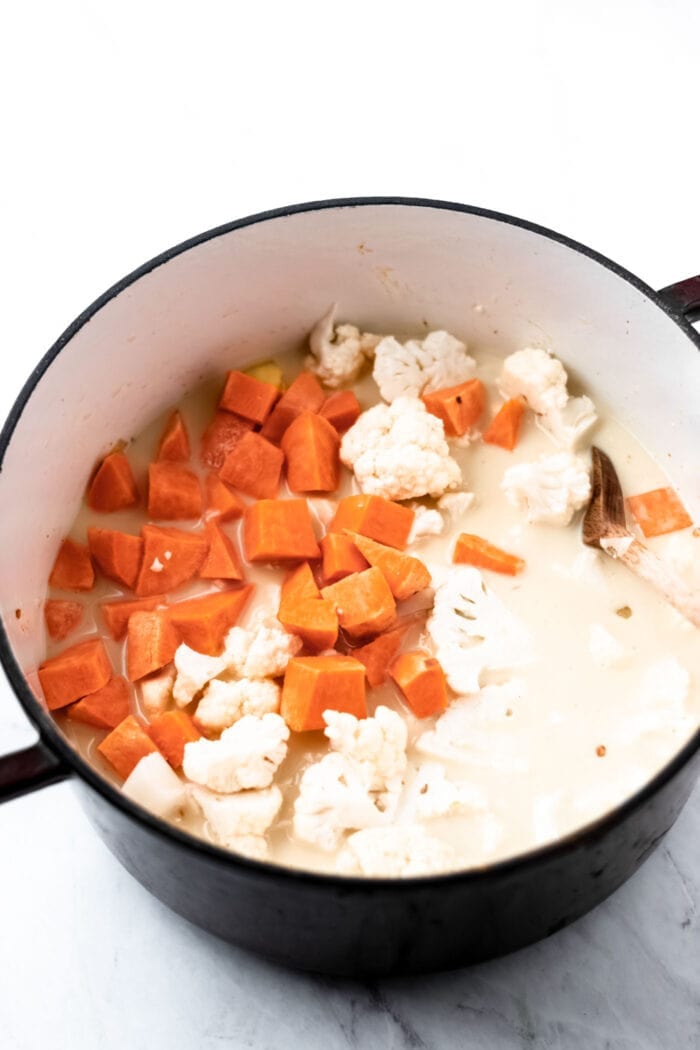 Chopped sweet potato, cauliflower and coconut milk being mixed in a large soup pot with a wooden spoon..