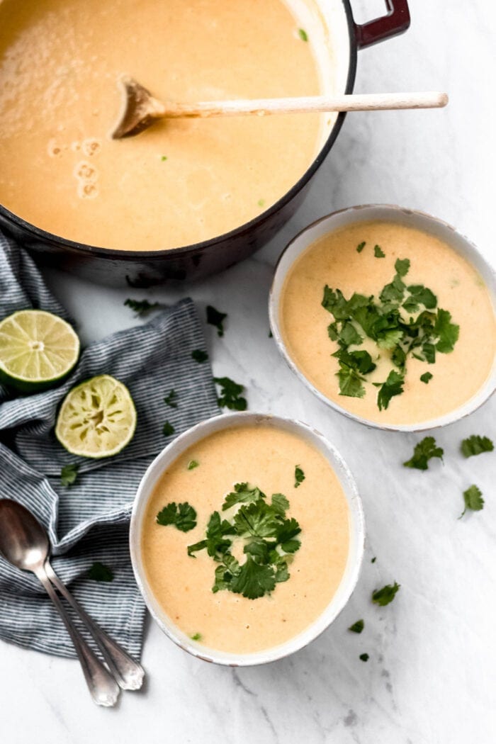 Two bowls and a large stock pot of cauliflower sweet potato soup with green peas. The bowls of soup are topped with fresh cilantro and there are some wedges of lime and two spoons beside them.